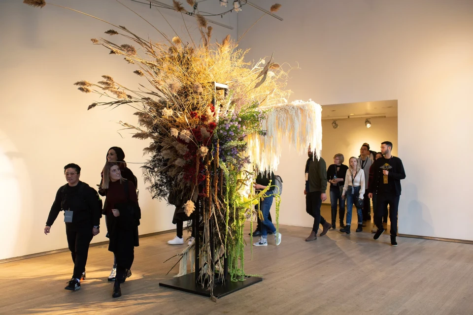 Large floral art installation with dried flowers and reeds at art gallery, and visitors walking by, adding life and movement to the scene.