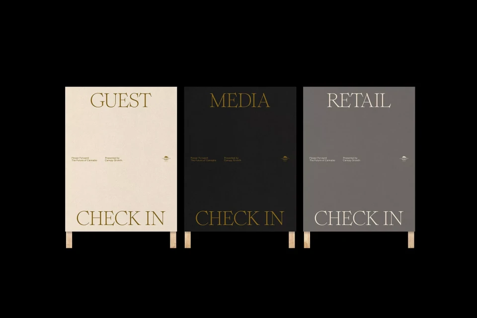 Three signs in black, white, and grey, on wood legs, read: Guest Check In, Media Check In, and Retail Check In on a black background.