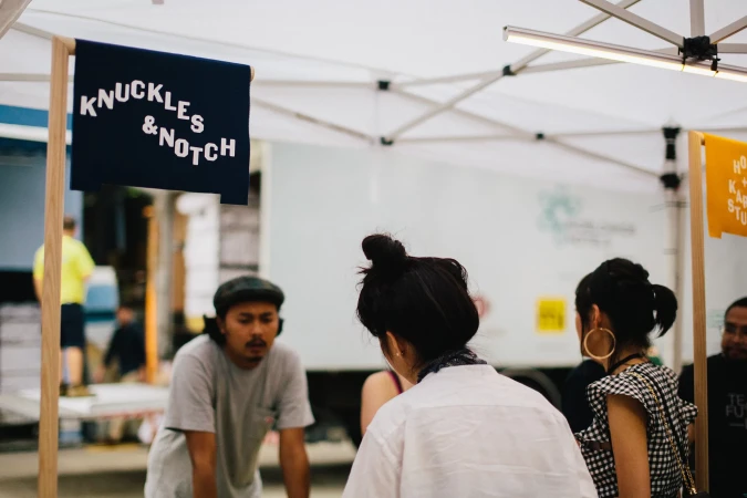 Outdoor marketplace scene with people browsing vendor booths under a white tent, featuring signs for local brands.