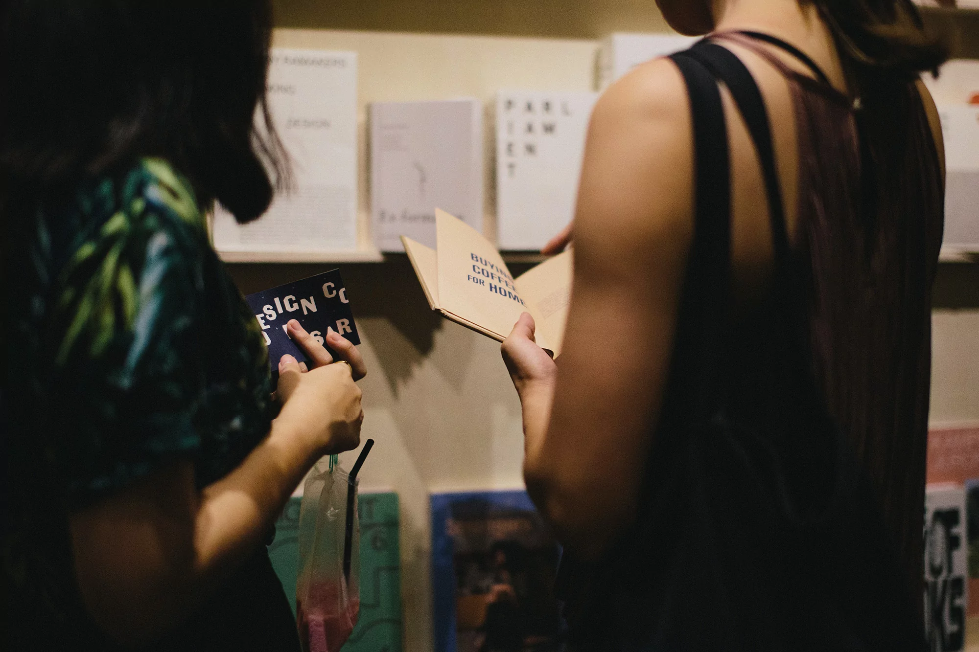 Two women browsing books at a bookstore. The woman on the right is reading a beige book while the other holds a dark book.