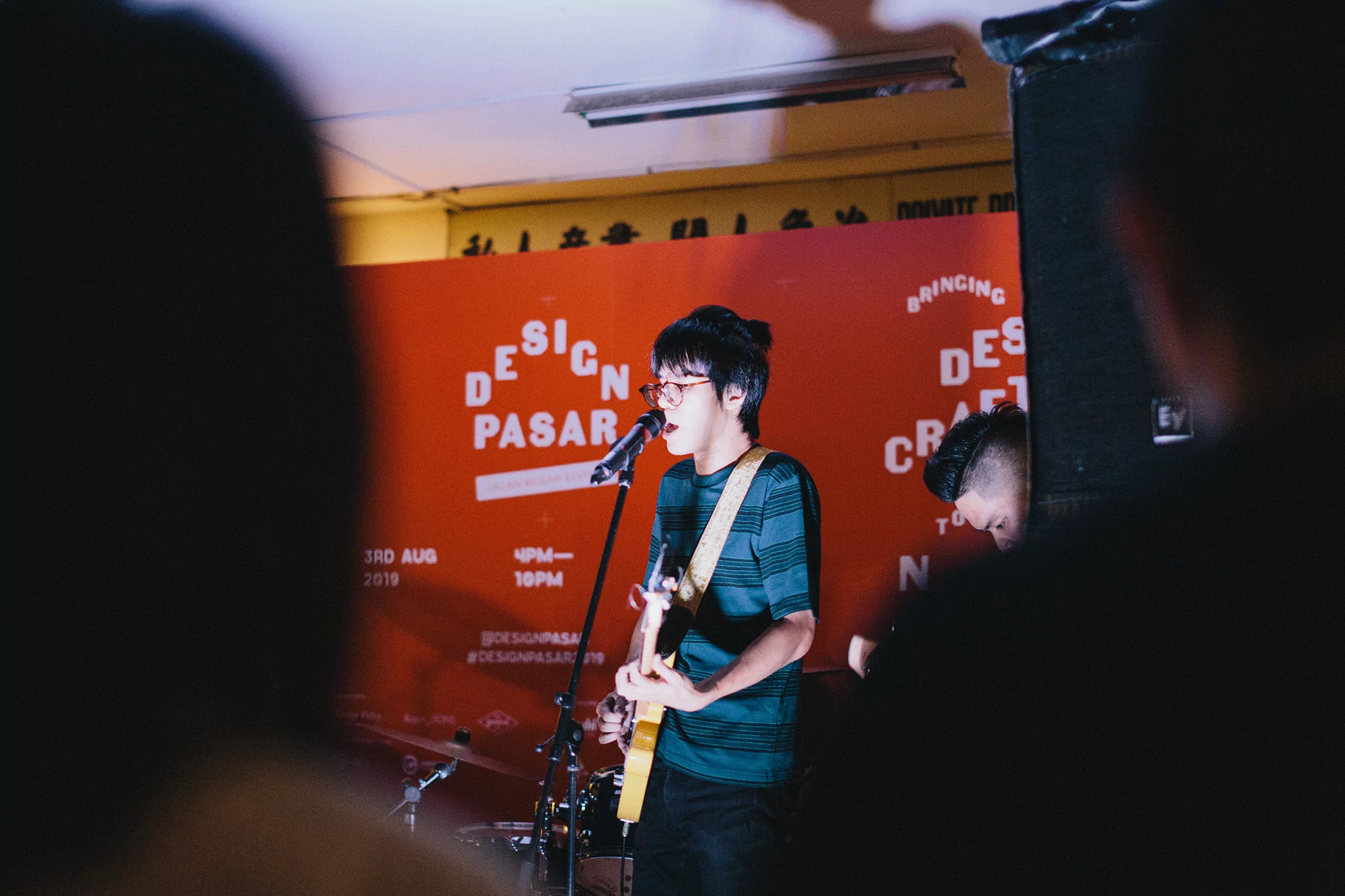A singer-guitarist performs on stage at an indoor music venue with an orange backdrop, captured from a low angle, partially obscured by silhouetted figures.