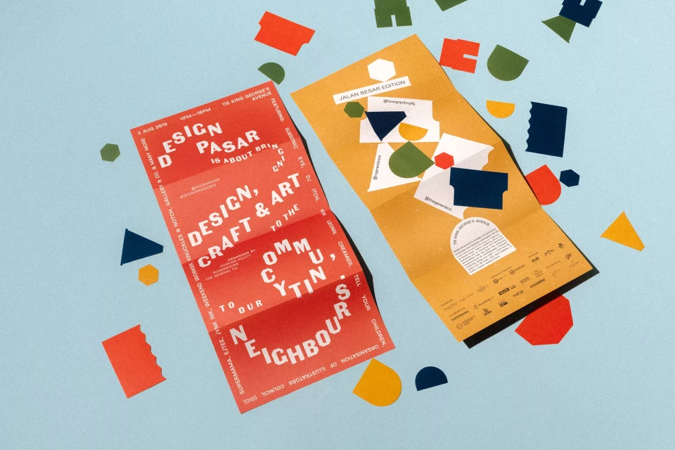 Two colorful, folded leaflets advertising a Design Pasar event, accented with geometric paper cutouts, lie on a light blue surface.