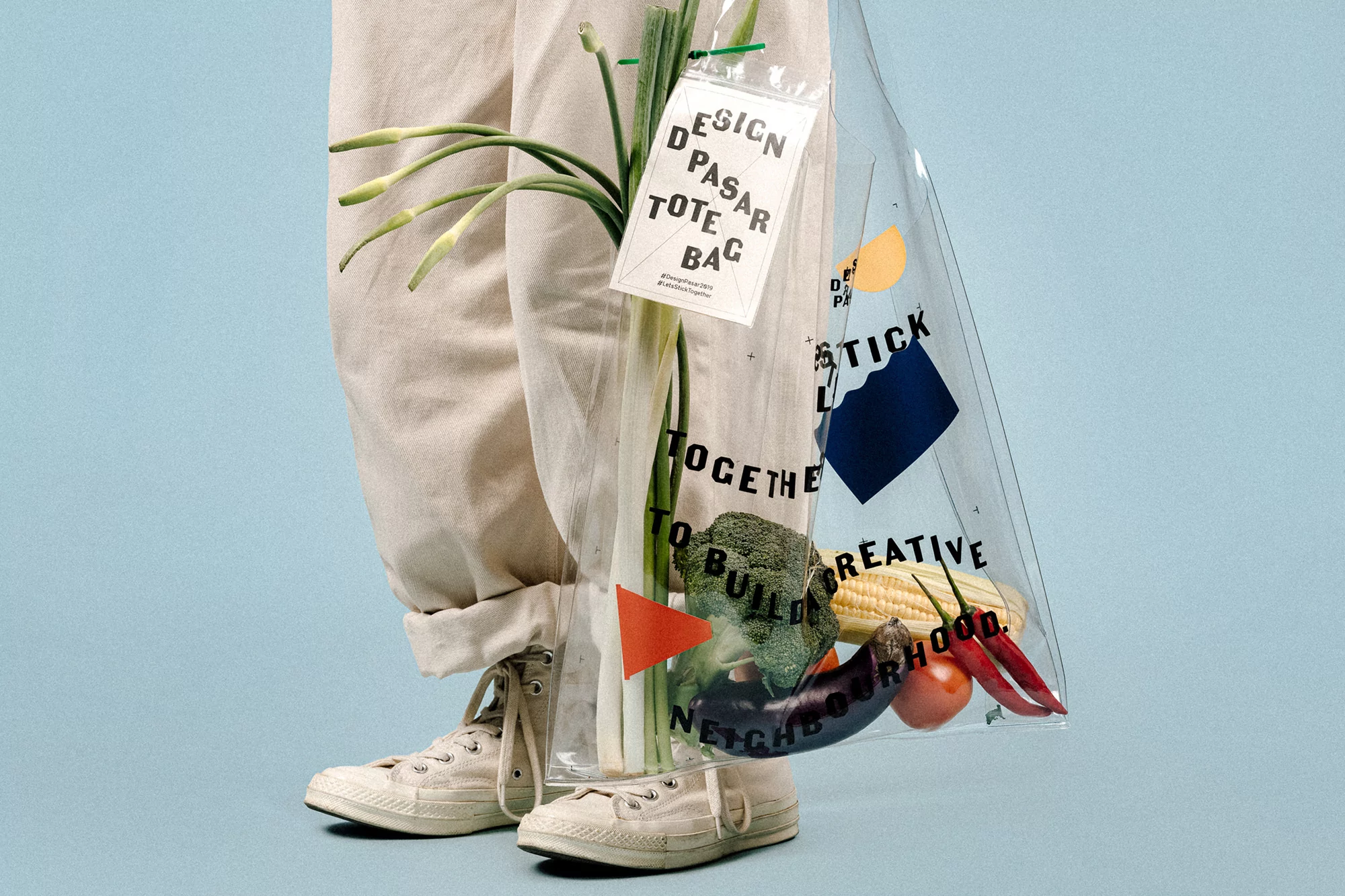 Person standing with a clear tote bag filled with vegetables, featuring a logo and text design, against a light blue background.