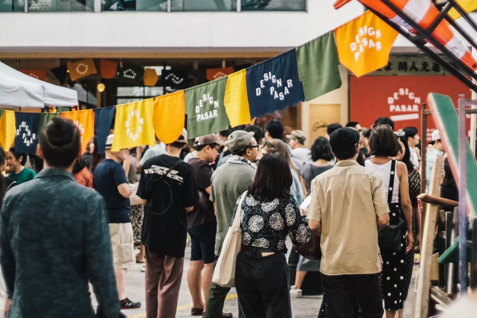 Outdoor market scene featuring many people, banners with text, and commercial activity in an urban setting during the daytime.
