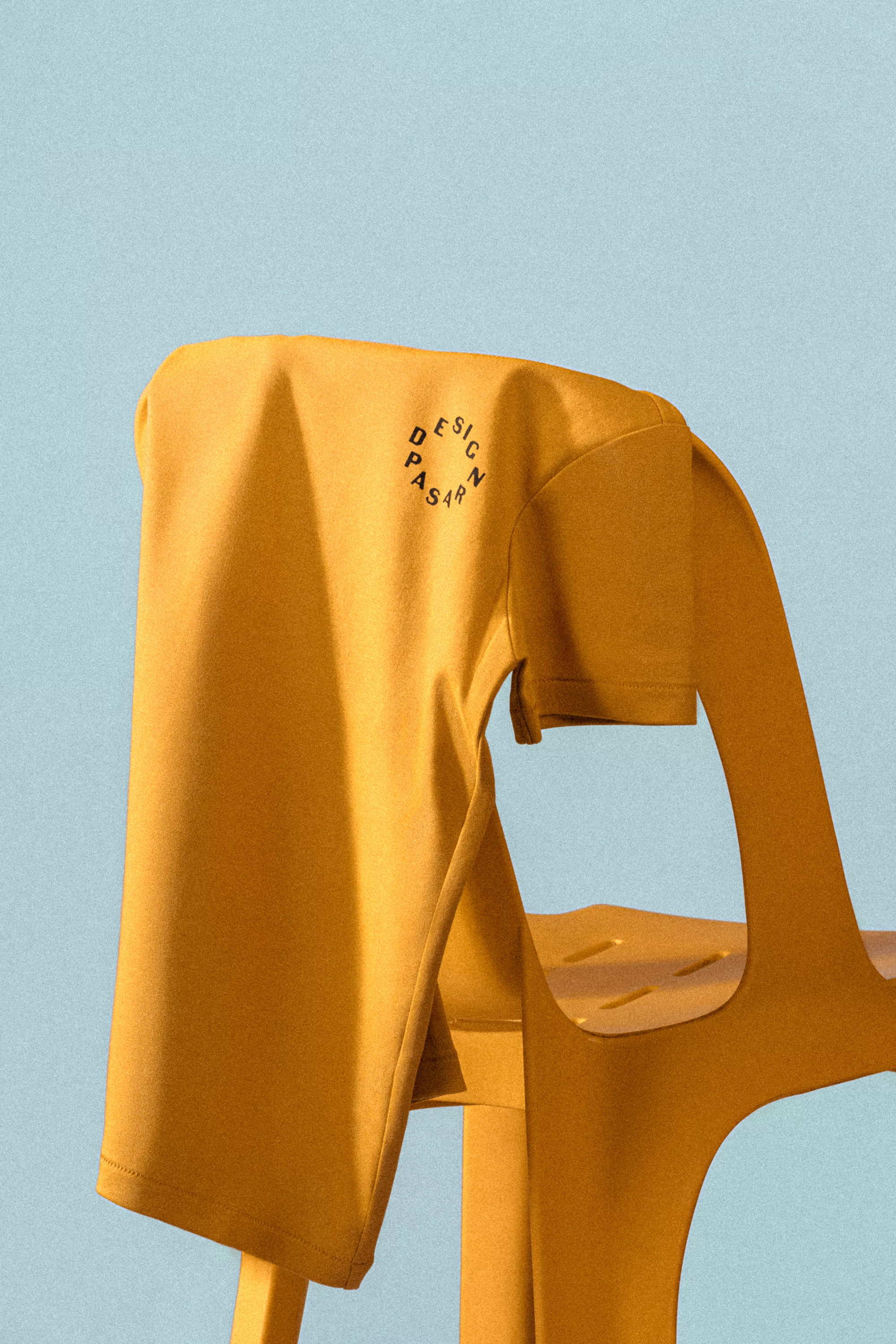 Mustard yellow T-shirt with a circular logo on a yellow chair against a light blue background in a studio shot.