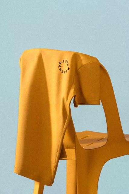 Mustard yellow T-shirt with a circular logo on a yellow chair against a light blue background in a studio shot.