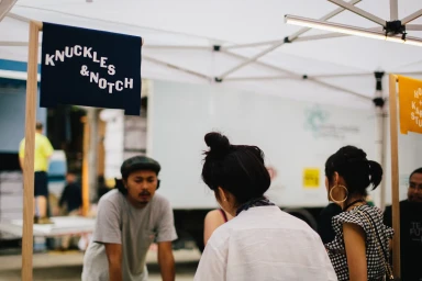 People gathered at an outdoor market stall with a sign reading Knuckles and Notch under a white canopy tent.