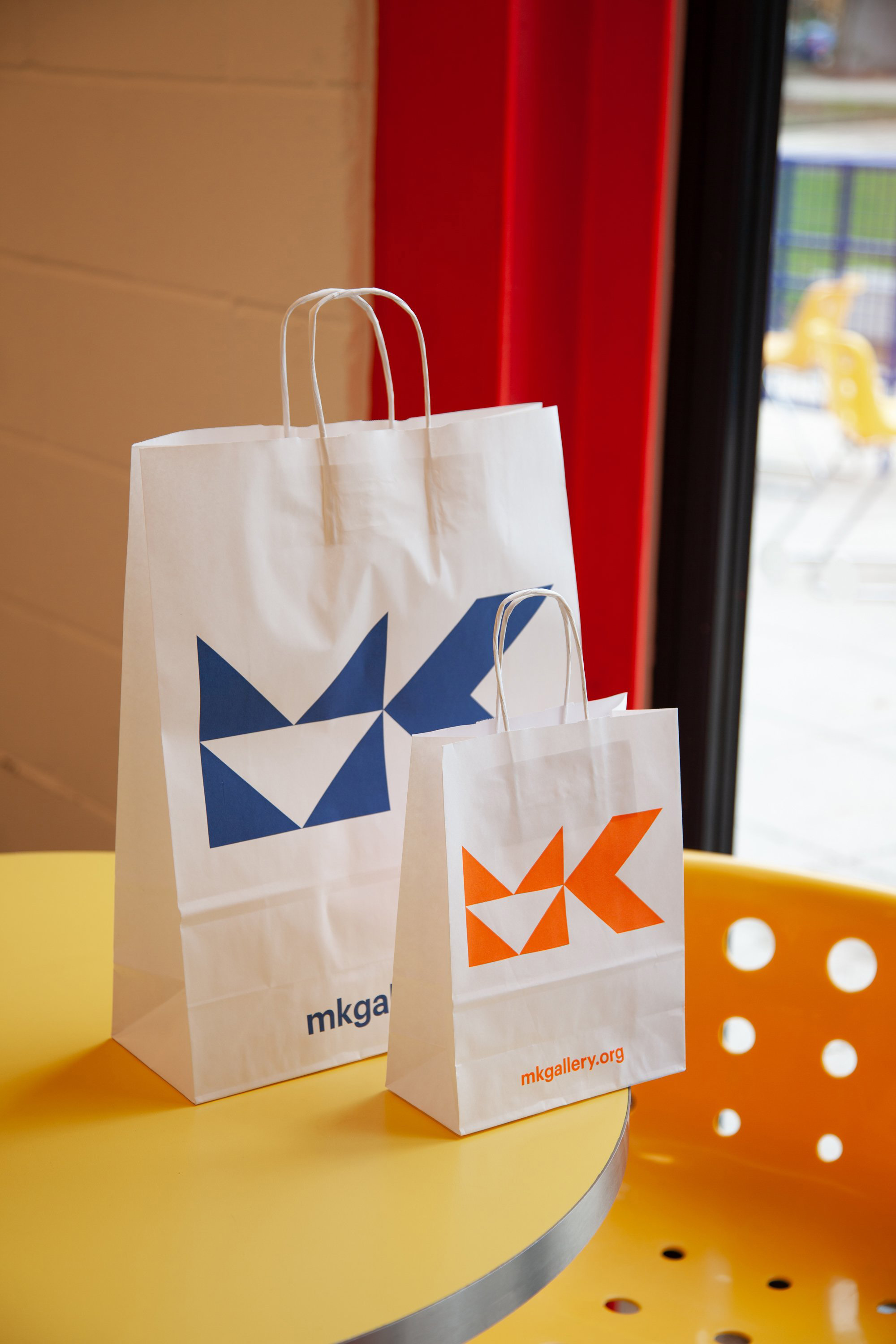 Two white paper shopping bags with geometric logo design on a yellow round table in an indoor setting with window and curtain.