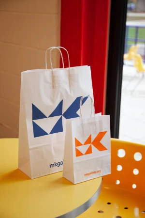Two white paper shopping bags with geometric logo design on a yellow round table in an indoor setting with window and curtain.