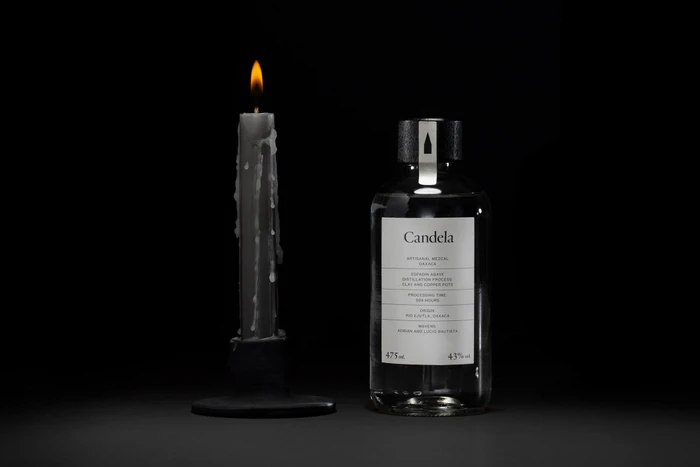 A lit dark candle next to a glass bottle of mezcal with a white label against a dark background