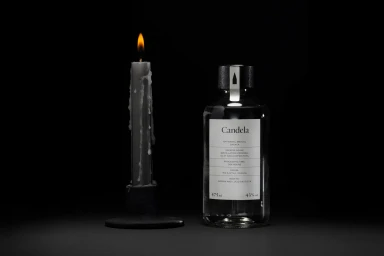 A lit dark candle next to a glass bottle of mezcal with a white label against a dark background