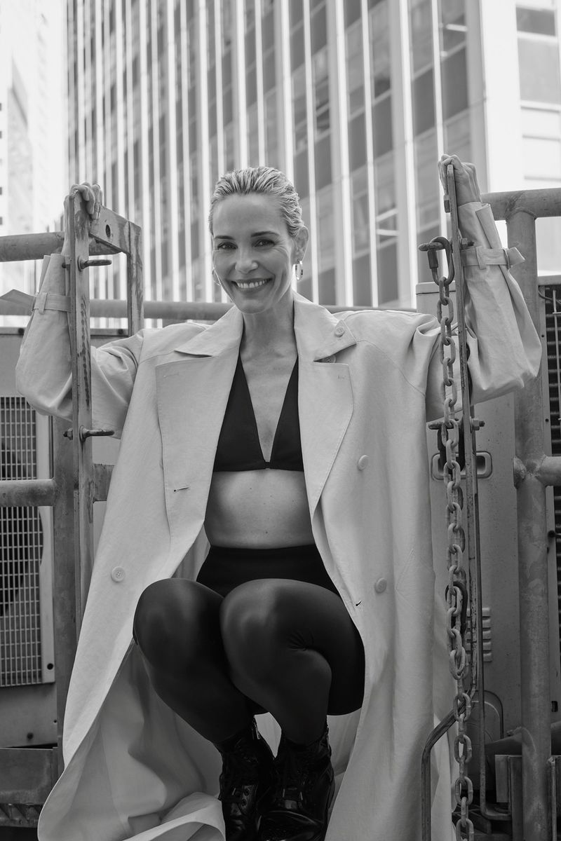 Stylish woman in bra top and trench coat poses on a building's scaffolding with city buildings in the background in black and white.