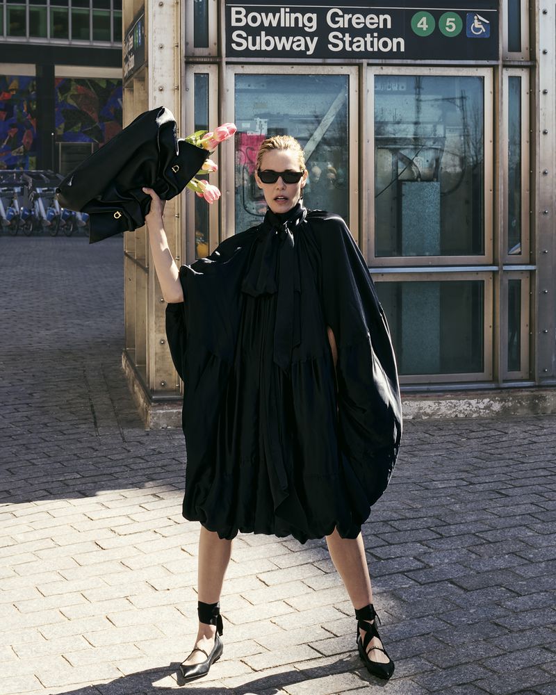 Woman in oversized black dress stands near subway entrance holding flowers and a jacket in an urban environment with bright sunlight.
