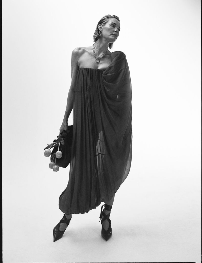Elegant woman in a flowing black dress and strappy heels poses with tulips against a white backdrop in a fashion-forward editorial shot.