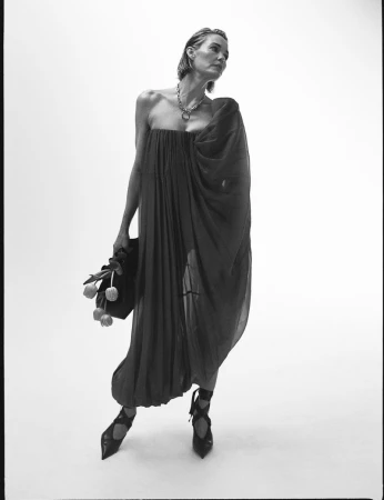 Elegant woman in a flowing black dress and strappy heels poses with tulips against a white backdrop in a fashion-forward editorial shot.