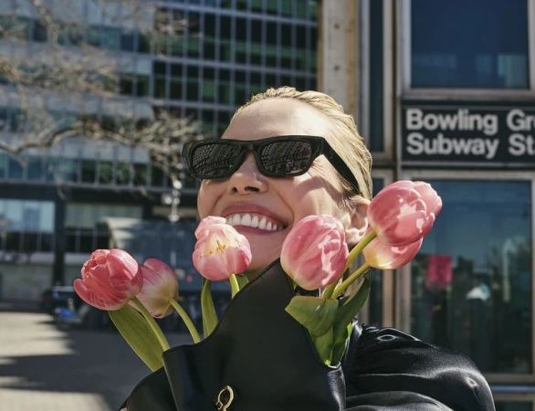 Smiling woman in sunglasses holds pink tulips in front of an urban building and subway sign, with a black purse visible.