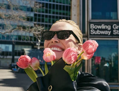 Leslie Bibb smiling outdoors while holding pink tulips and wearing black sunglasses and a black garment.