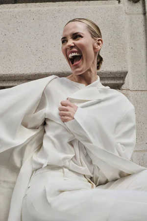 Leslie Bibb laughing wearing a white outfit against a stone architectural background