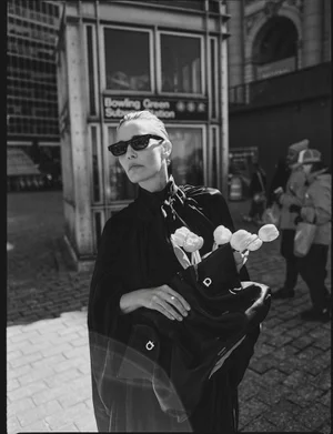 Leslie Bibb in dark sunglasses wearing a black top and holding a black handbag with white tulips against an urban street background.