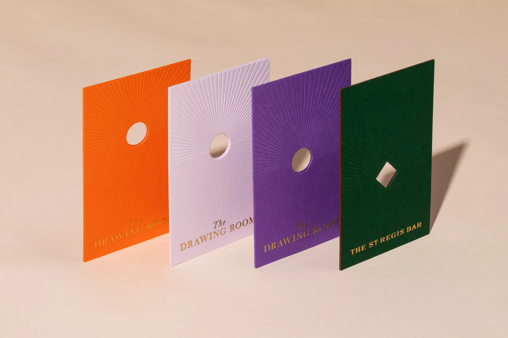 Four textured paper cards in orange, lilac, purple, and green with geometric designs, each displaying elegant gold lettering.