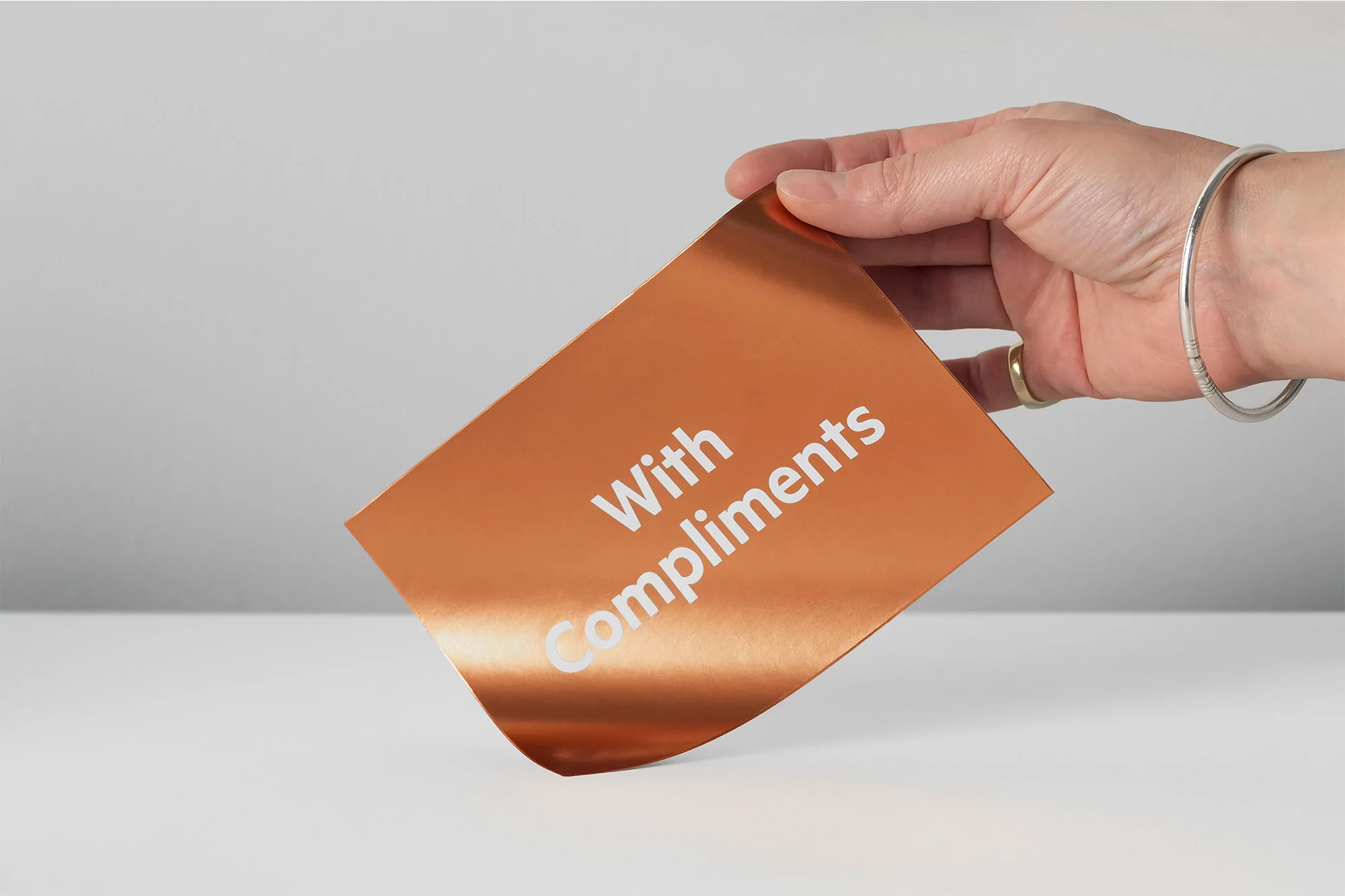 Hand holding a copper-colored card with the text 'With Compliments' against a light gray background, showcasing a promotional message.