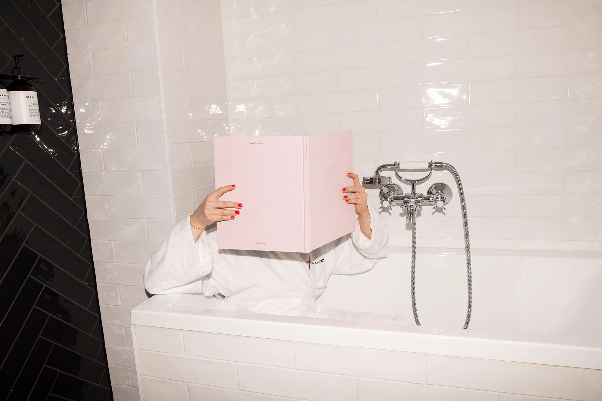 Person in bathtub wearing a white robe, holding a pink book in front of their face, with white and black tile in the background.