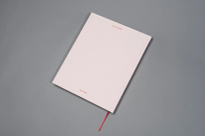 Closed light pink hardcover book with a red ribbon bookmark resting on a neutral grey surface.