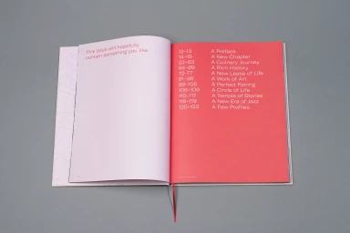 Open book with a pink page on the left and a bright red table of contents page on the right resting on a grey surface.