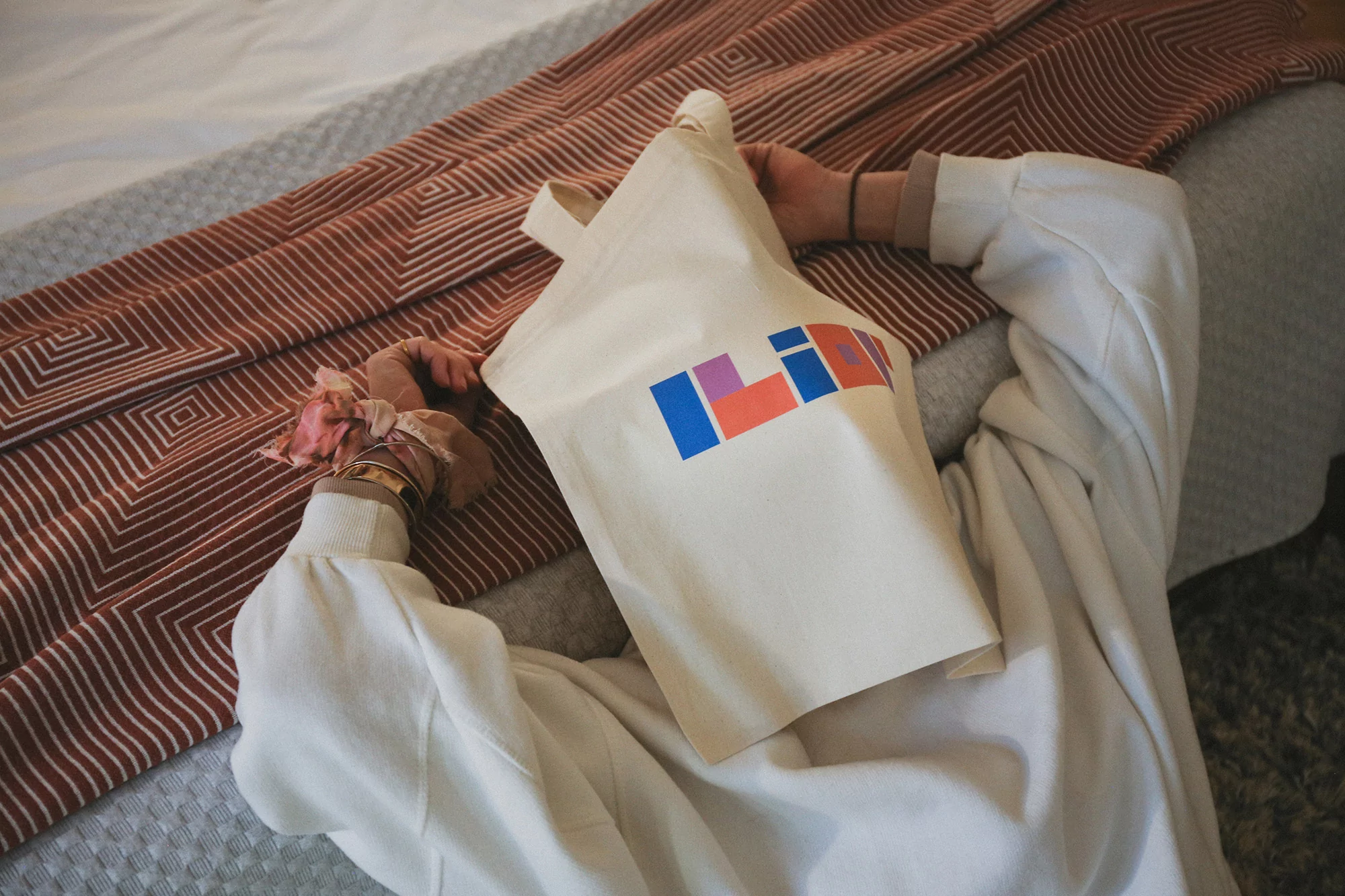 A person lying on a bed with a cream canvas tote bag featuring a colourful Ilior logo covering their face, wearing a white sweater.