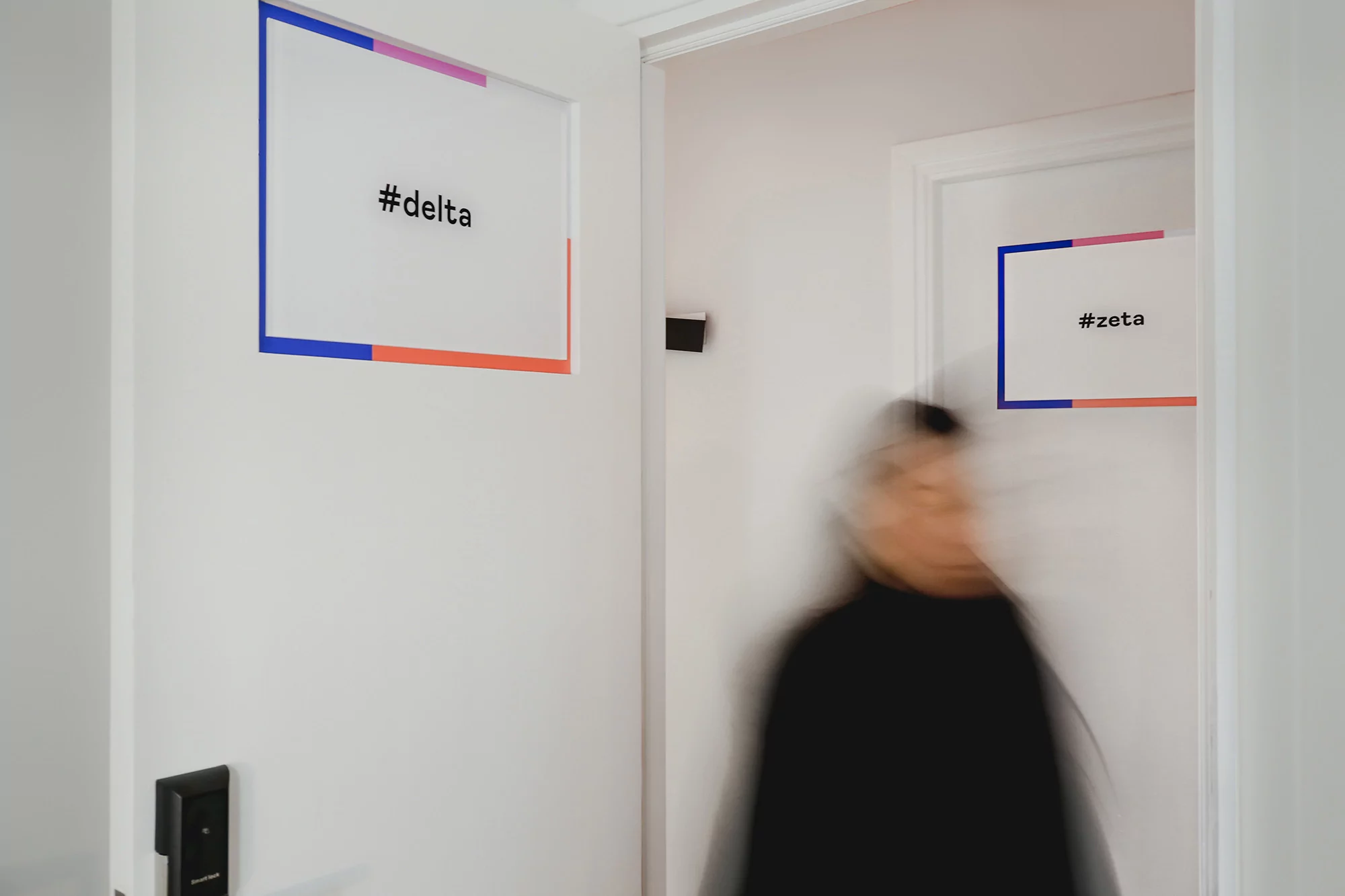 Blurred figure walks past white doors labeled '#delta' and '#zeta', creating a modern, minimal interior scene with colorful accents.
