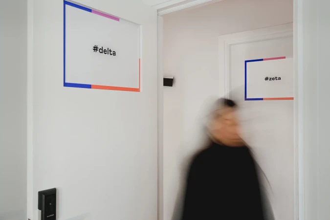 Blurred figure walks past white doors labeled '#delta' and '#zeta', creating a modern, minimal interior scene with colorful accents.