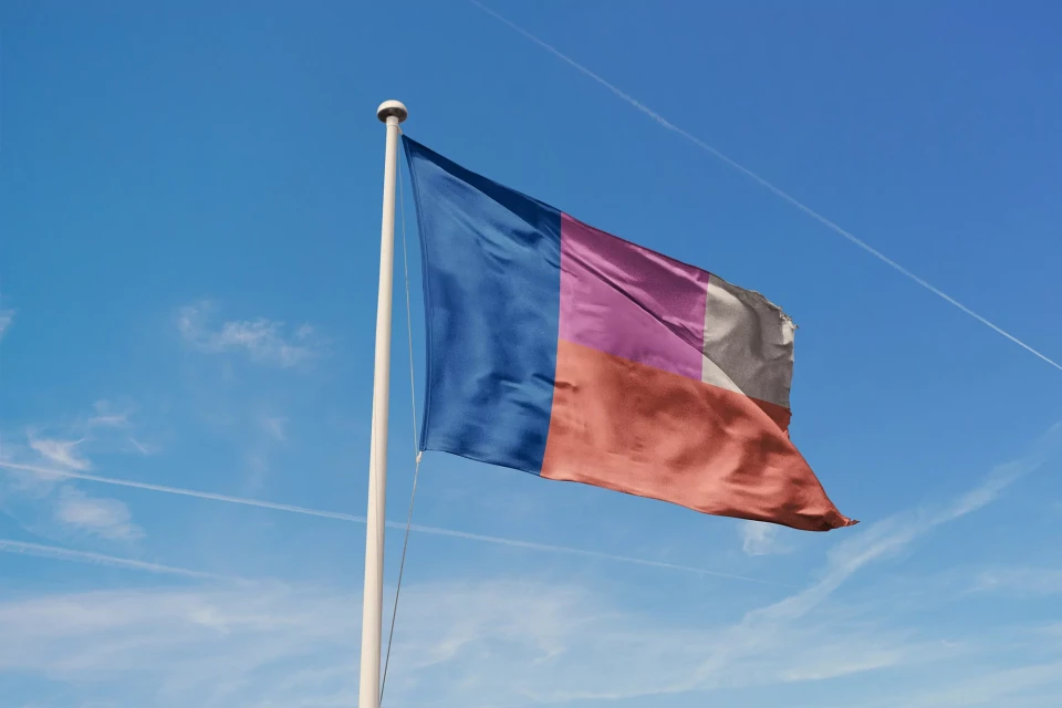 A colorful flag with a blue, pink, brown and grey color block design waving against a bright blue sky with contrails visible above.