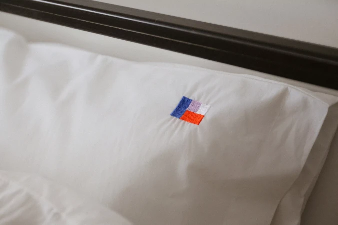 Close-up of a white pillow on a bed featuring a small embroidered geometric multi-colored logo patch by Ilior.