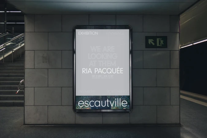 Billboard promoting 'Ria Pacquée' art exhibit displayed on wall with staircase in background in indoor location with neutral lighting.
