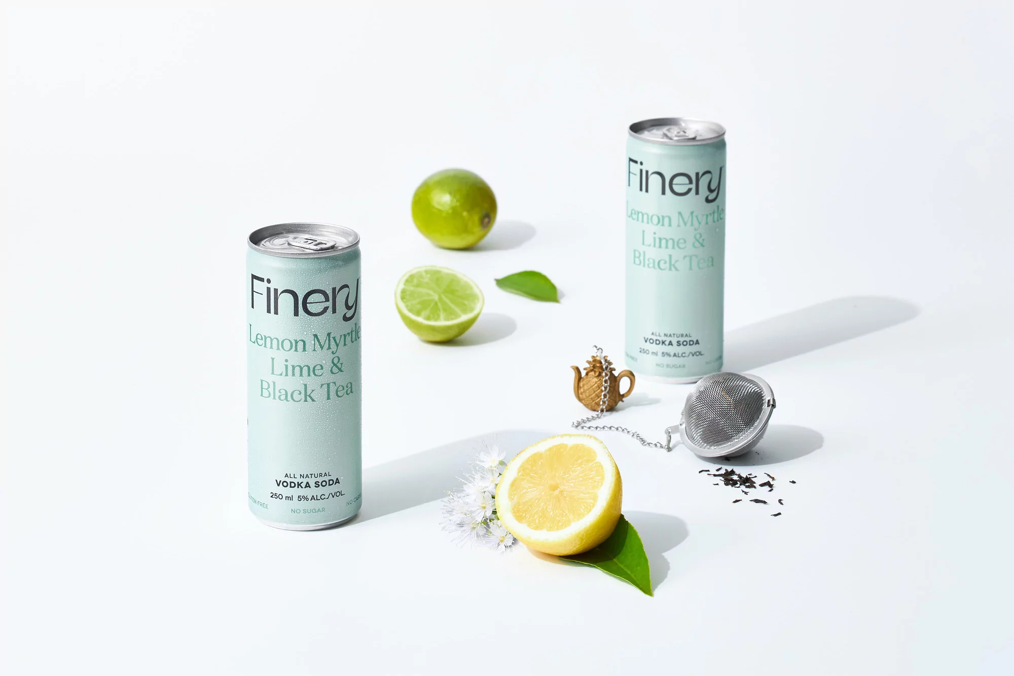 Two cans of Finery Lemon Myrtle Lime & Black Tea soda, limes, lemon half, tea infuser, and tea on a white surface in a studio shot.