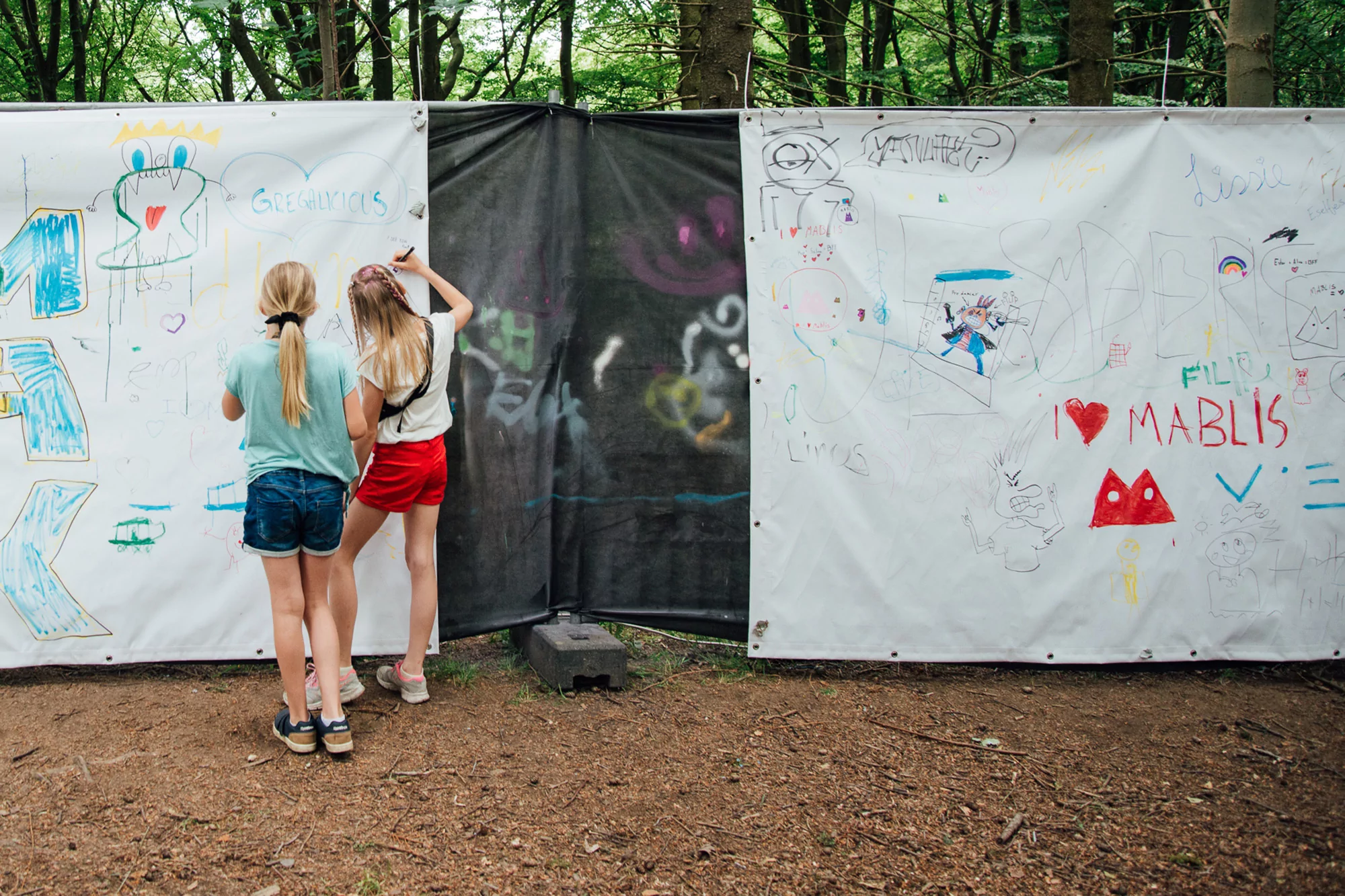 Two children draw on a white banner in a wooded area. One child is drawing with a marker, with scribbles and colorful artwork.