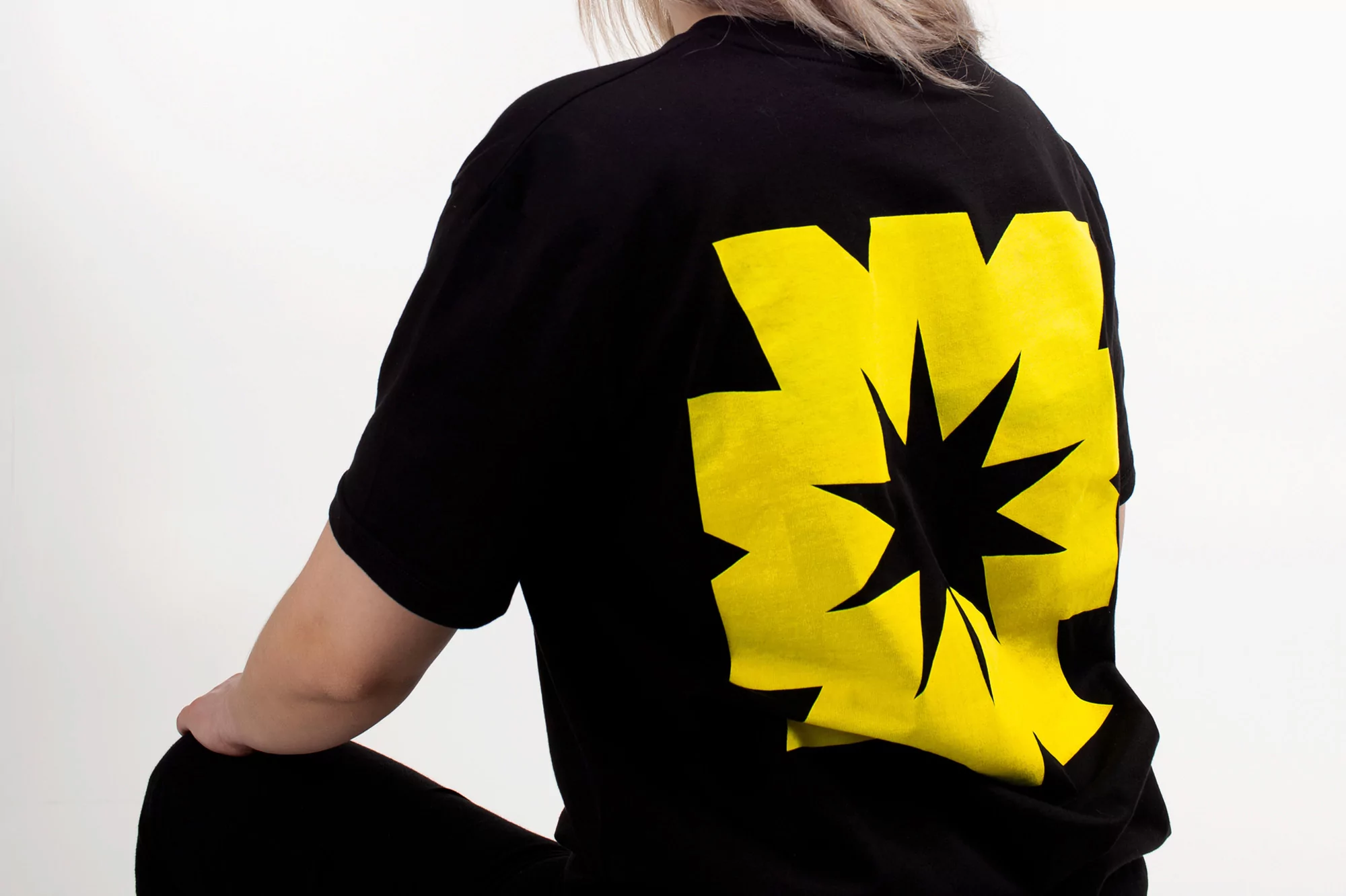 Blonde woman wears a black t-shirt with a yellow star graphic on the back, posing against a white backdrop in a studio setting.