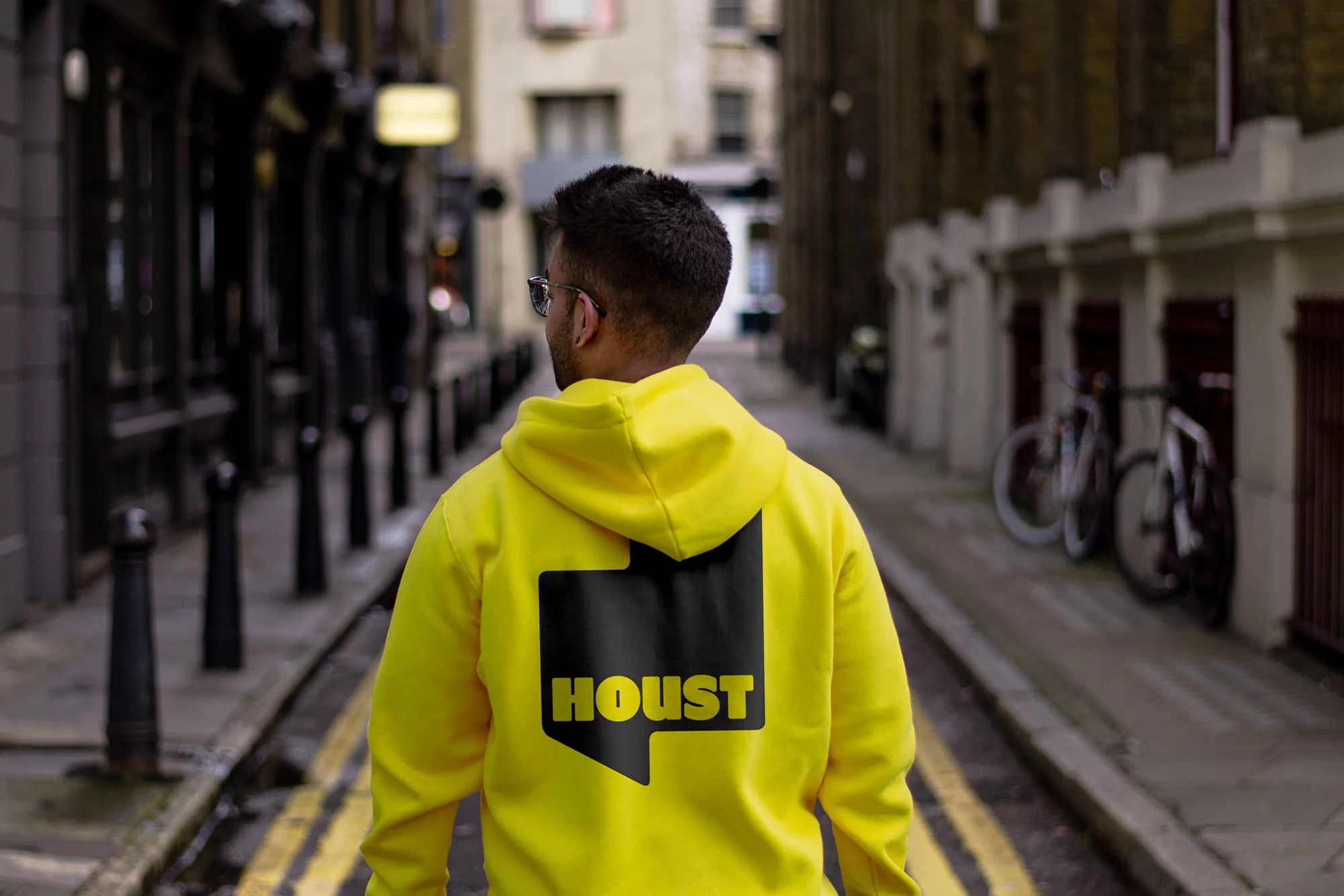Back view of a stylish person in a bright yellow hoodie walking down a narrow urban street lined with bollards and buildings.