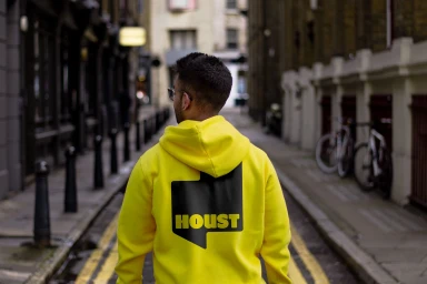 Man wearing a bright yellow hoodie with a black Houst logo walking down a narrow urban street