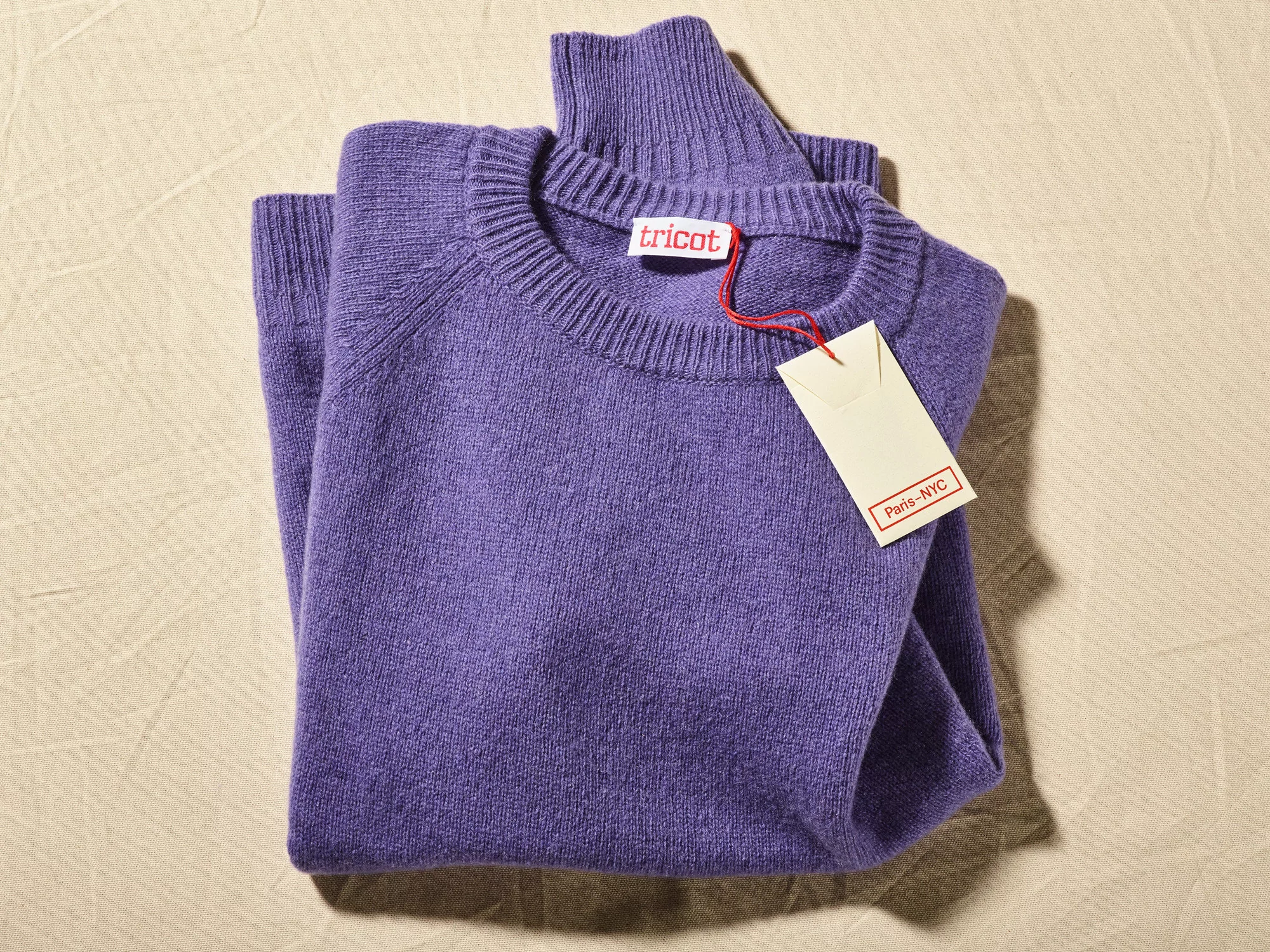 Folded purple knit sweater with a white tag on a textured beige background. Soft lighting and natural fabric texture.
