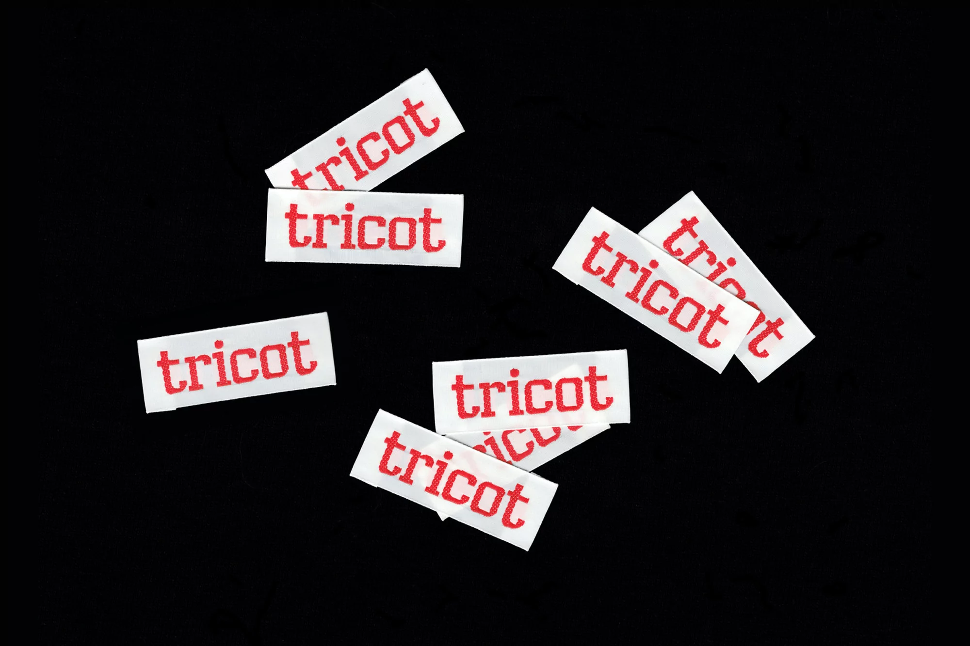 Collection of woven clothing labels against black background with 'tricot' in red lettering. Tags are white and arranged at different angles.