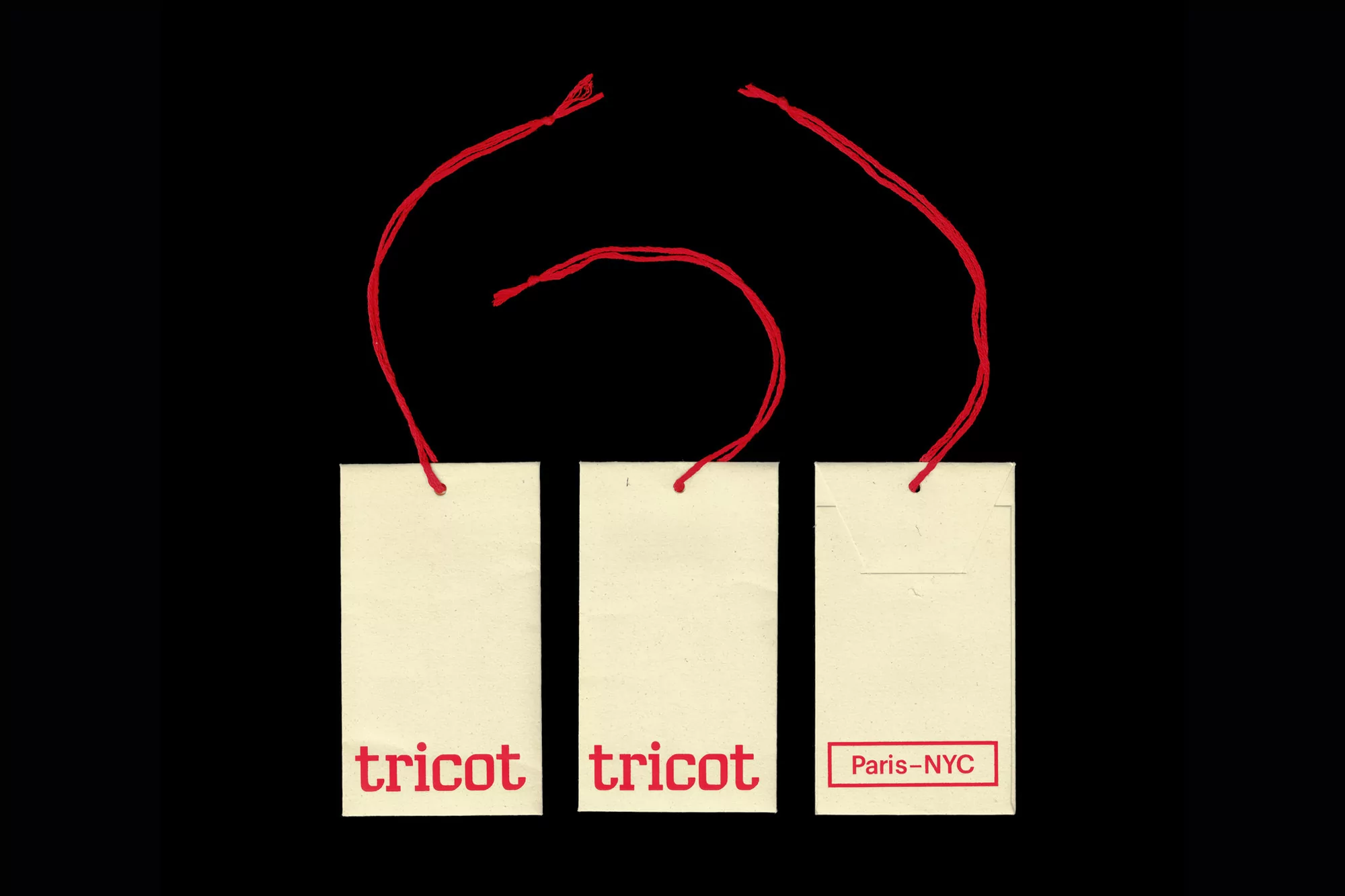 Three cream-colored tags with red string and red text, 'tricot' and 'Paris-NYC' against a stark black background, product tags, fashion