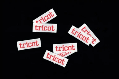 Scattered rectangular white paper labels featuring red tricot typography against a solid black background.