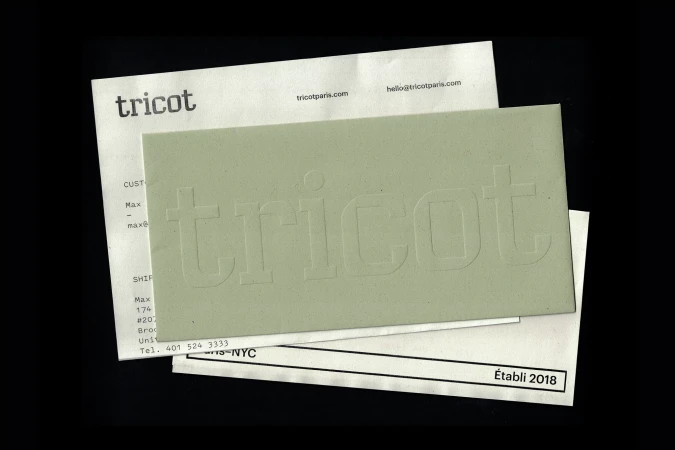 Overhead view of Tricot brand stationery featuring an embossed sage green paper envelope resting on white printed documents against a black ground