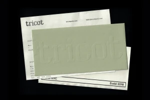 Overhead view of Tricot brand stationery featuring an embossed sage green paper envelope resting on white printed documents against a black ground