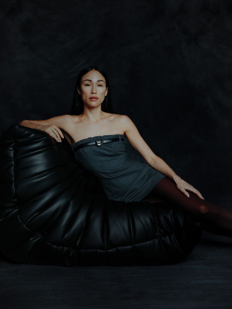 A woman reclines on a black leather beanbag chair against a dark background, wearing a grey strapless dress and reddish-brown tights.