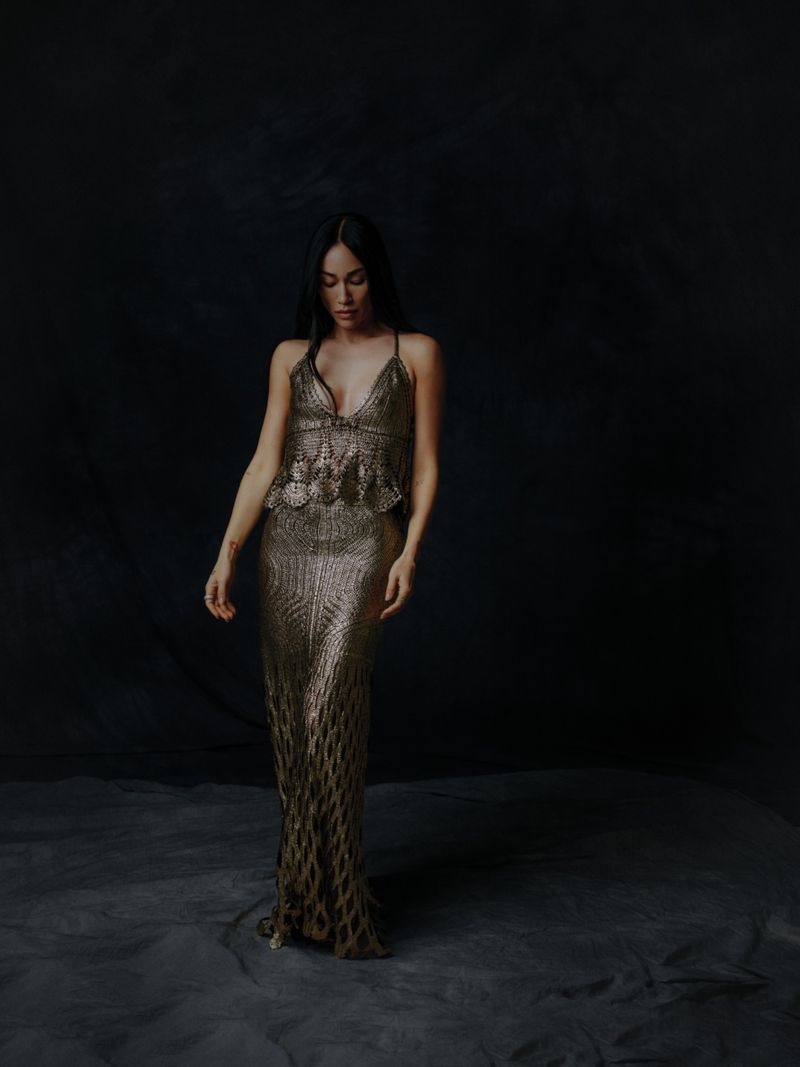 A full-length portrait captures a woman with long, dark hair wearing a gold, shimmering gown against a dark, textured backdrop.