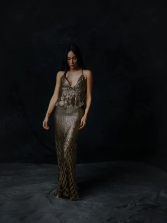A full-length portrait captures a woman with long, dark hair wearing a gold, shimmering gown against a dark, textured backdrop.