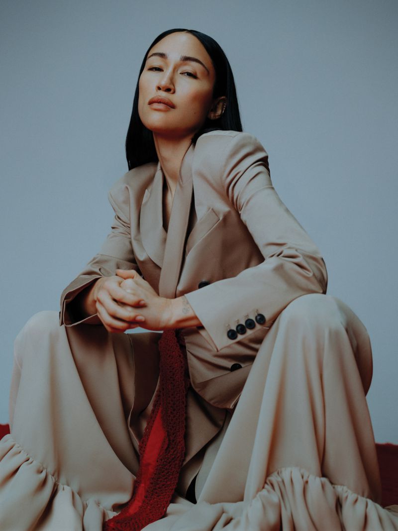 Elegant woman in a beige blazer and pants ensemble, seated indoors against a light grey background, gazing slightly upward.