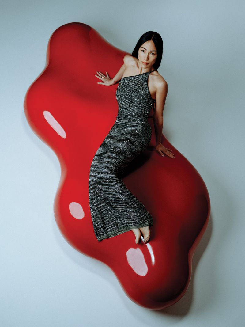 Adult woman in silver dress poses on a large red lip-shaped modern sofa in a minimalist indoor studio setting.